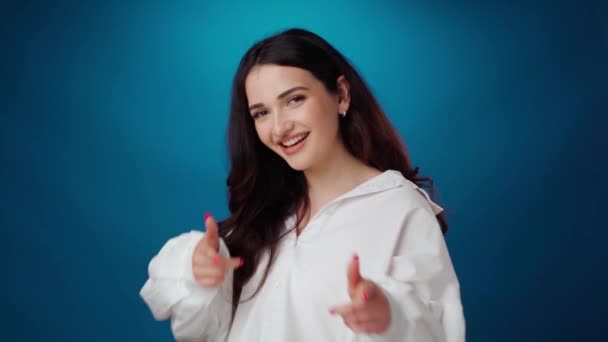 Freundlich lächelnde junge Frau zeigt vor blauem Hintergrund in die Kamera — Stockvideo