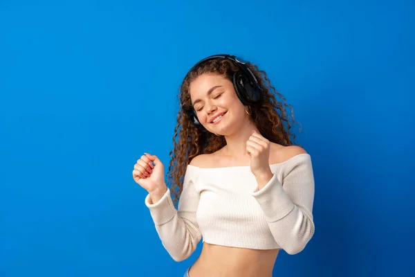 stock image Young attractive curly woman listening to music with headphones against blue background