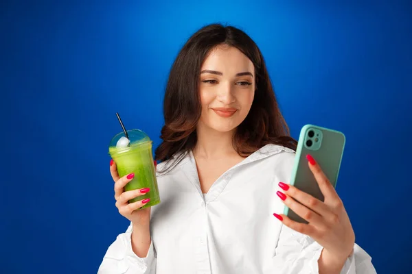 Giovane donna felice attraente in possesso di una tazza di frullato verde e utilizzando il suo smartphone su sfondo blu — Foto Stock