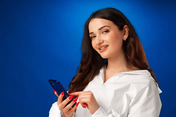 Sorrindo jovem mulher em camisa branca usando seu smartphone contra fundo azul — Fotografia de Stock