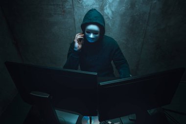Yüz maskesi takan anonim hacker karanlık odada bilgisayarla çalışıyor.