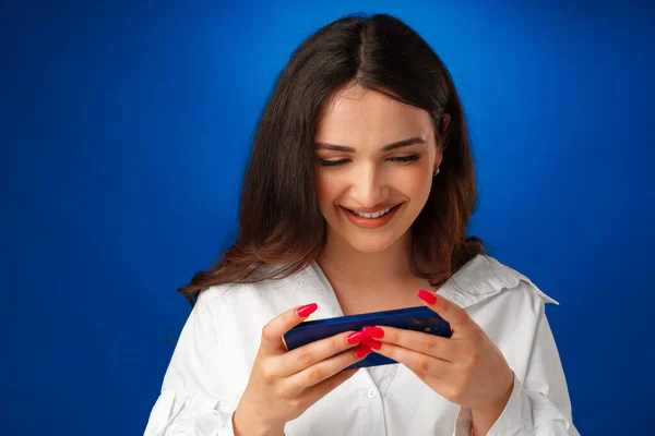 Sorrindo jovem mulher em camisa branca usando seu smartphone contra fundo azul — Fotografia de Stock