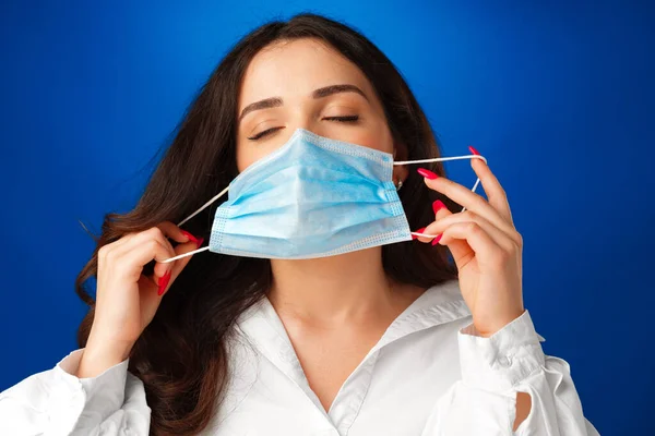 Молодая женщина снимает медицинскую маску с лица на синем фоне — стоковое фото