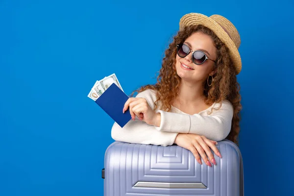 Młoda piękna kobieta w kapeluszu na wakacjach z walizką na niebieskim tle — Zdjęcie stockowe