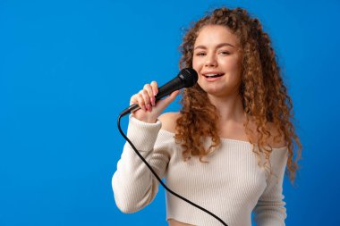 Genç, kıvırcık saçlı, mavi arka planda mikrofonda şarkı söyleyen kadın.