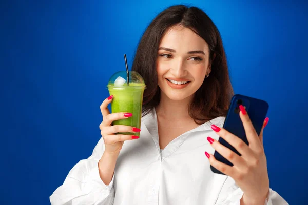 Jovem mulher feliz atraente segurando uma xícara de smoothie verde e usando seu smartphone contra fundo azul — Fotografia de Stock