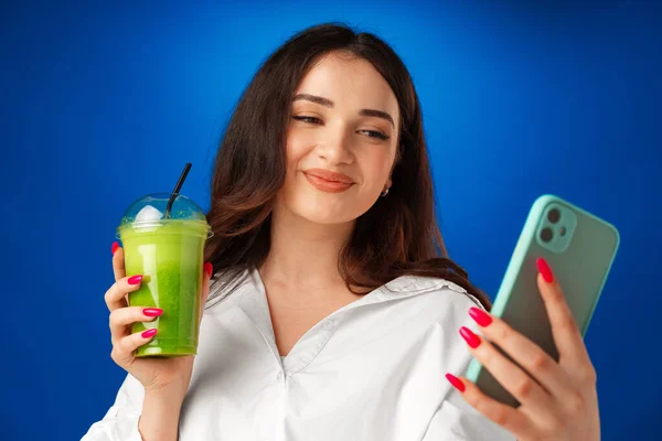 Jovem mulher feliz atraente segurando uma xícara de smoothie verde e usando seu smartphone contra fundo azul — Fotografia de Stock