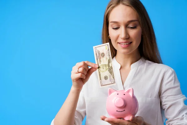 Mujer poniendo dinero en la alcancía rosa contra fondo azul — Foto de Stock