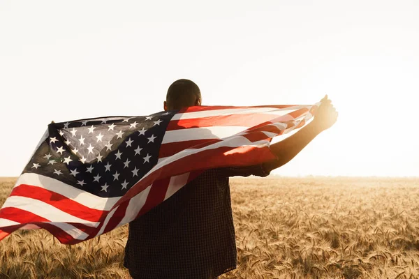 Young man holding American flag on back while standing in wheat field — Stock Photo, Image