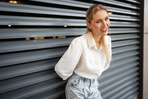 Stijlvol meisje in jeans en witte katoenen blouse poseren in de buurt van grijze muur — Stockfoto