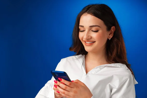 Sorrindo jovem mulher em camisa branca usando seu smartphone contra fundo azul — Fotografia de Stock