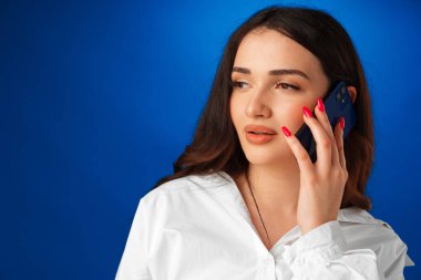 Genç esmer kadın telefonda mavi arka planda konuşuyor.