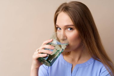 Güzel, sağlıklı genç bir kadın bej arka planda bir bardaktan su içiyor.