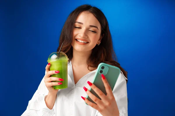 Jeune femme heureuse attrayant tenant une tasse de smoothie vert et en utilisant son smartphone sur fond bleu — Photo