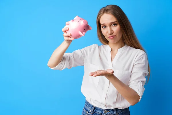 Nešťastná žena drží prasátko banka peníze box proti modrému pozadí — Stock fotografie
