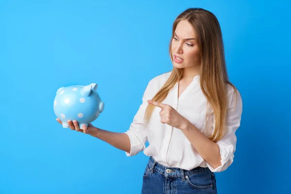 Mujer infeliz sosteniendo alcancía moneybox contra fondo azul — Foto de Stock