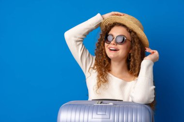 Mavi arka planda bavuluyla gezen şapkalı güzel bir kadın.