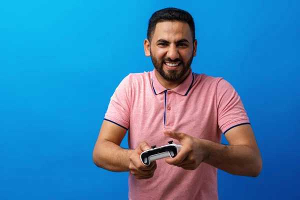 Pembe tişörtlü genç Arap adam mavi arka planda video oyunları oynuyor. — Stok fotoğraf