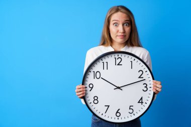 Çekici genç bir kadın büyük duvar saatini mavi arka planda tutuyor.