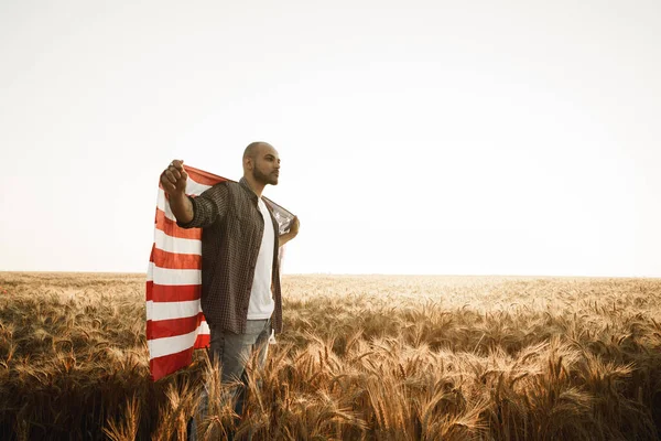 Joven afroamericano sosteniendo bandera nacional de Estados Unidos a través del campo de trigo —  Fotos de Stock