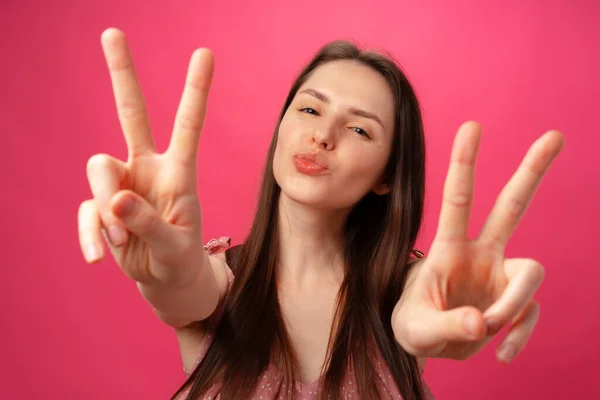 Junge lächelnde brünette Frau zeigt Siegeszeichen vor rosa Hintergrund — Stockfoto