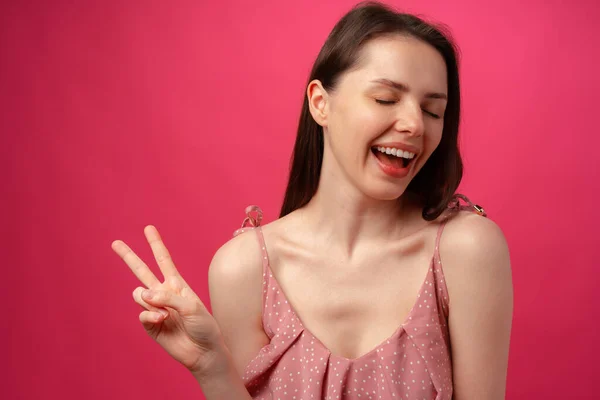 Junge lächelnde brünette Frau zeigt Siegeszeichen vor rosa Hintergrund — Stockfoto