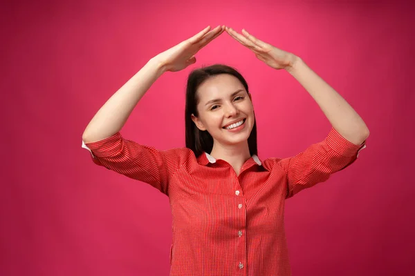 Young brunette woman holding hands above head like roof against pink background