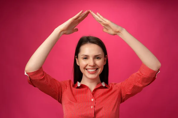 Junge brünette Frau hält die Hände über dem Kopf wie ein Dach vor rosa Hintergrund — Stockfoto