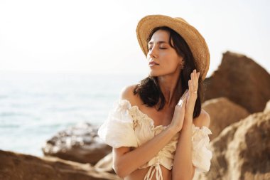 Genç, neşeli esmer kadın deniz ve kayalara karşı poz veriyor.