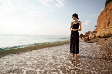 Genç, neşeli esmer kadın deniz ve kayalara karşı poz veriyor.