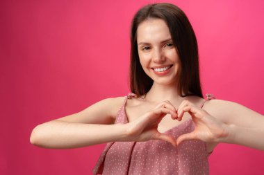 Gülümseyen genç bir kadın kalp işareti yapıyor. Elleriyle sevgisini gösteriyor.