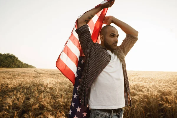 Joven afroamericano sosteniendo bandera nacional de Estados Unidos a través del campo de trigo —  Fotos de Stock