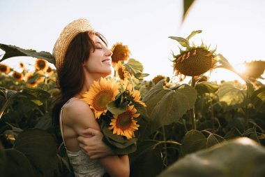 Gün batımında ayçiçeği tarlasında duran şapkalı, gülümseyen bir kadın.