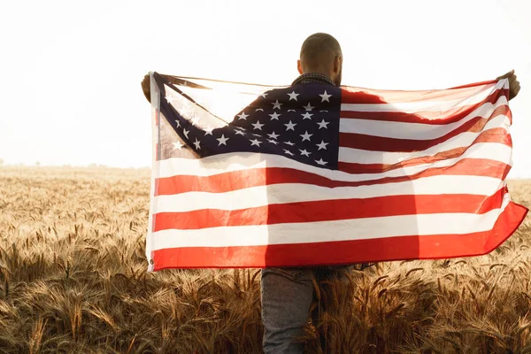 Joven sosteniendo bandera americana en la espalda mientras está de pie en el campo de trigo —  Fotos de Stock