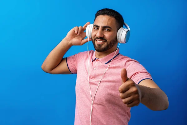 Jeune homme arabe heureux avec écouteurs écoutant de la musique sur fond bleu — Photo