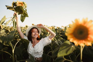 Gün batımında ayçiçeği tarlasında duran şapkalı, gülümseyen bir kadın.