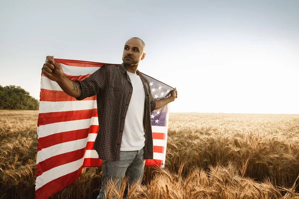 Jovem afro-americano segurando bandeira nacional dos EUA através do campo de trigo — Fotografia de Stock