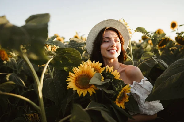 석양에 해바라기 밭에서 있는 모자를 쓴 미소짓는 여자 — 스톡 사진
