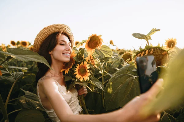Молода усміхнена жінка в капелюсі фотографує селфі в соняшниковому полі — стокове фото