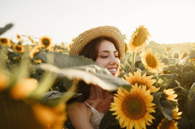 Gün batımında ayçiçeği tarlasında duran şapkalı, gülümseyen bir kadın.