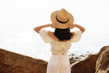 Genç, neşeli esmer kadın deniz ve kayalara karşı poz veriyor.