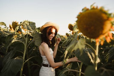 Gün batımında ayçiçeği tarlasında duran şapkalı, gülümseyen bir kadın.