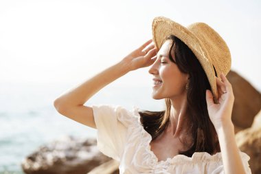 Genç, neşeli esmer kadın deniz ve kayalara karşı poz veriyor.