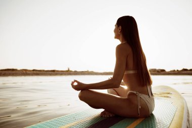 Woman practicing yoga on the paddle board in the morning clipart