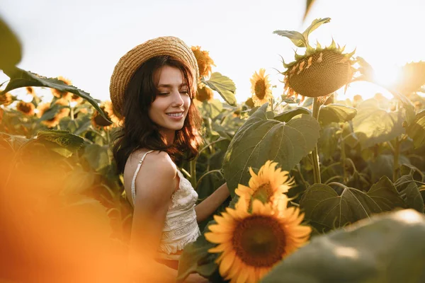 Усміхнена жінка в капелюсі стоїть на полі соняшників на заході сонця — стокове фото