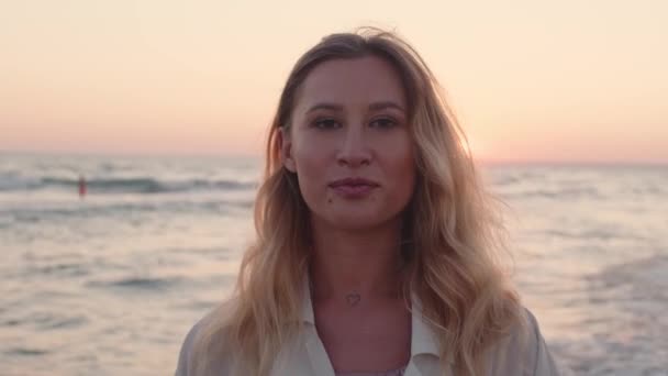 Jeune femme blonde aux cheveux ondulés debout sur la plage au bord de la mer au coucher du soleil — Video