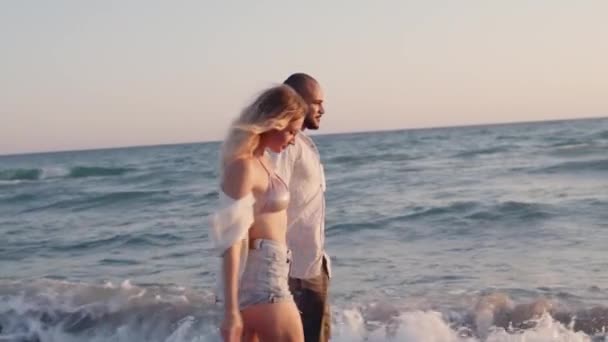 Пара молодых влюбленных, держащихся за руки, прогуливаясь по пляжу у моря — стоковое видео