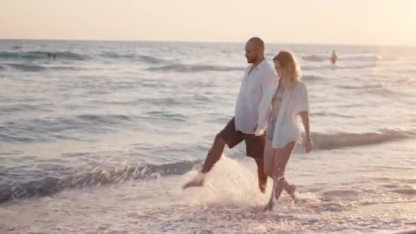 Пара молодых влюбленных, держащихся за руки, прогуливаясь по пляжу у моря — стоковое видео