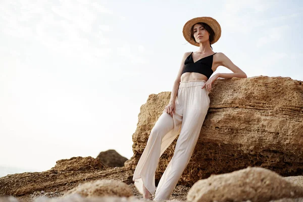 Retrato de mujer en bralette negro y pantalón blanco apoyado en roca en la playa — Foto de Stock