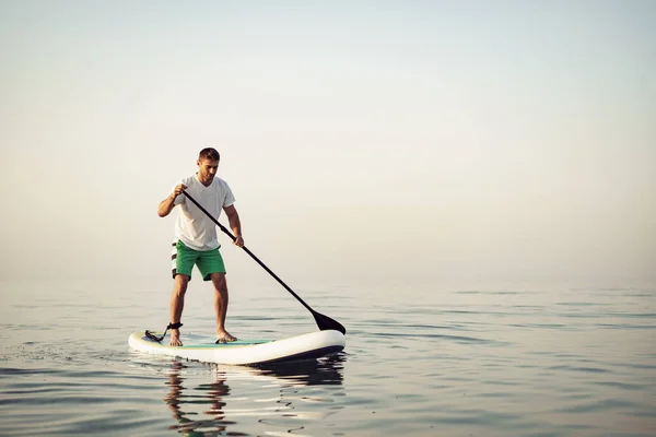 Jongeman in t-shirt en korte broek drijvend op SUP board — Stockfoto